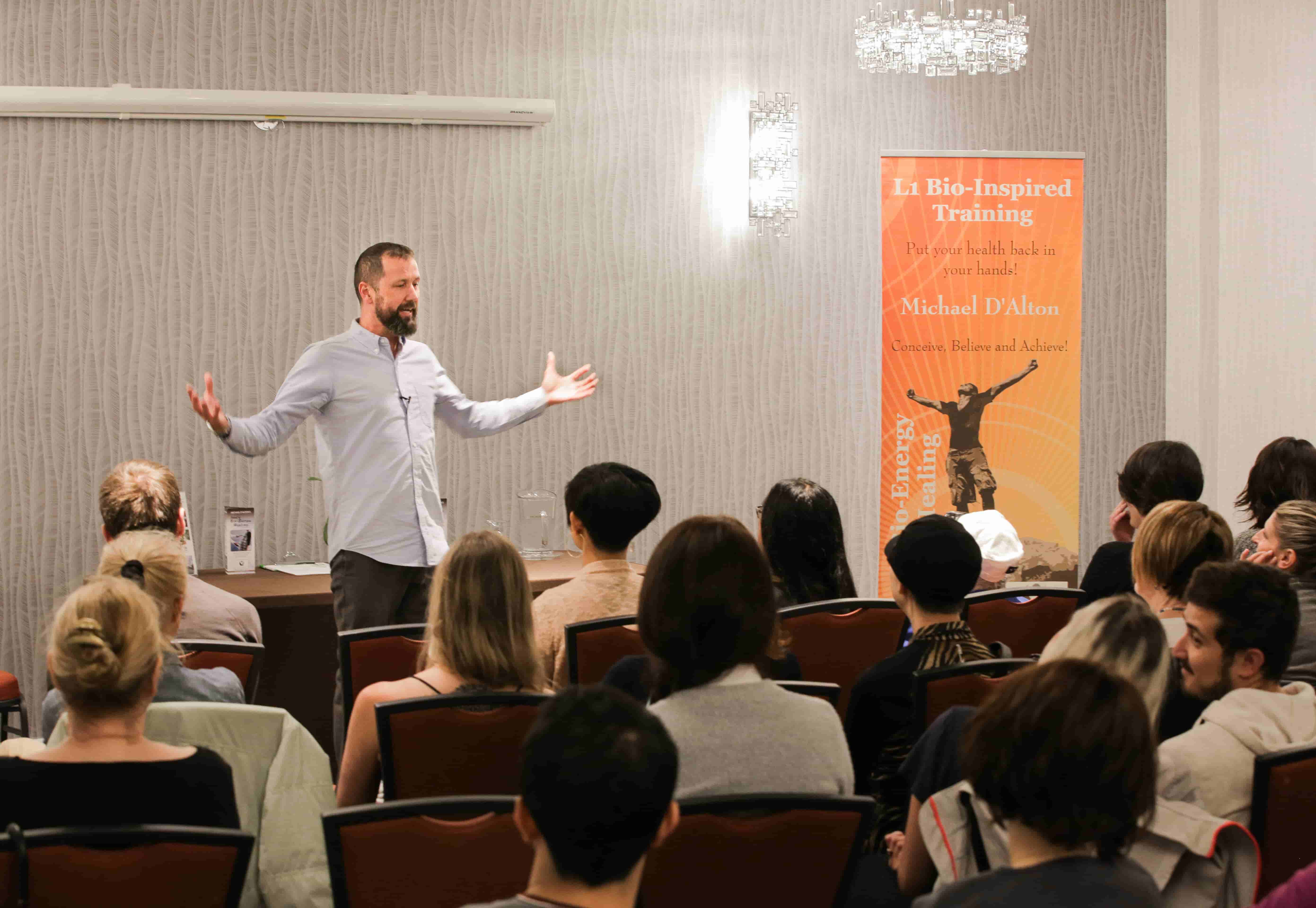 A man addresses a seated audience with his arms outstretched beside a Level 1 training banner.