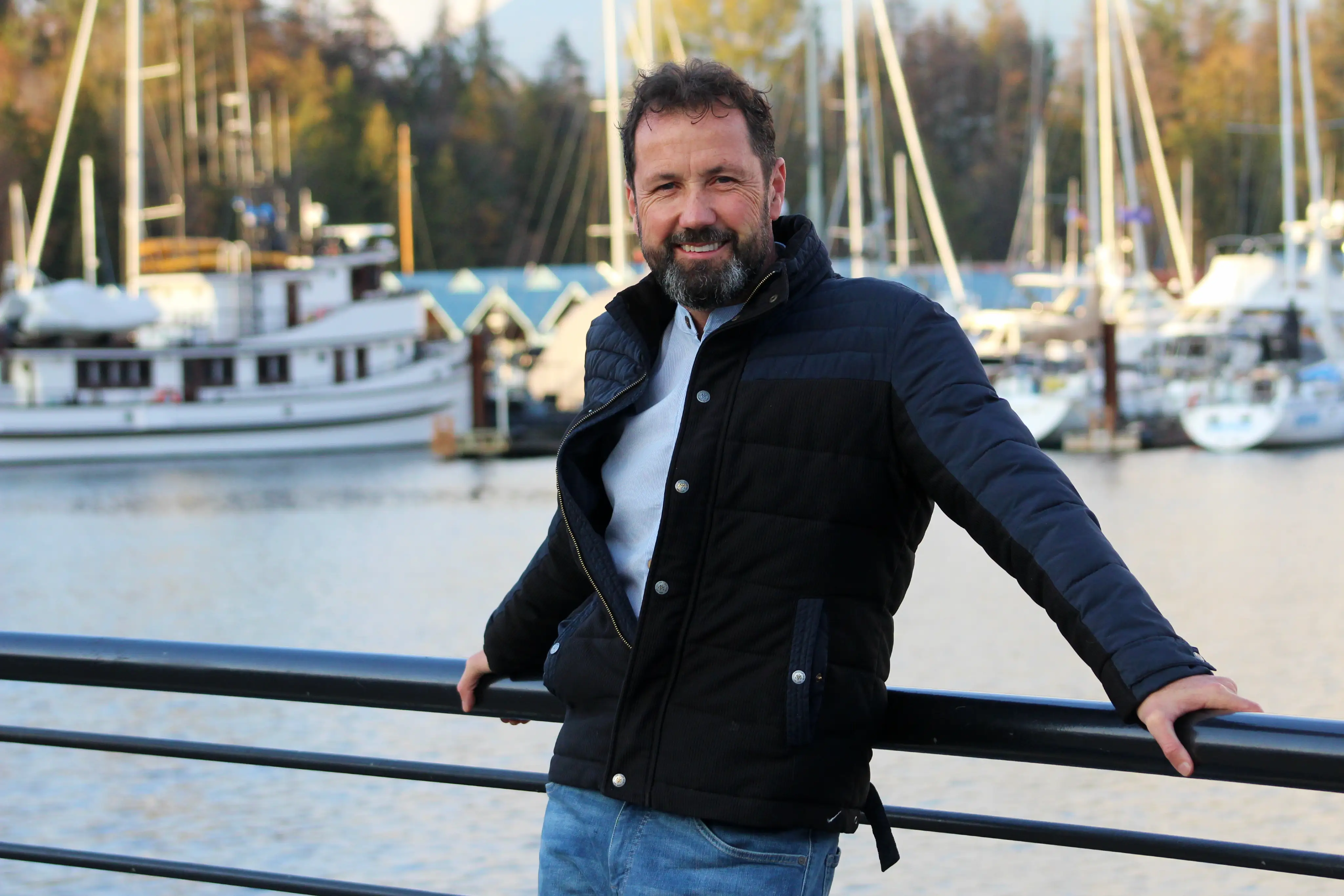A smiling man leans against a railing with boats moored behind him.